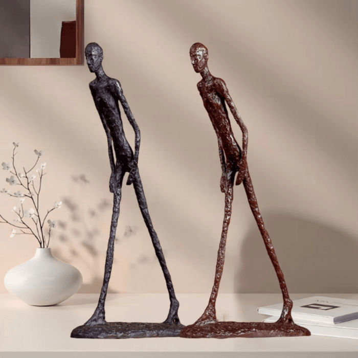 Two bronze Giacometti-inspired walking man sculptures with elongated limbs on a shelf with decor elements in the background.