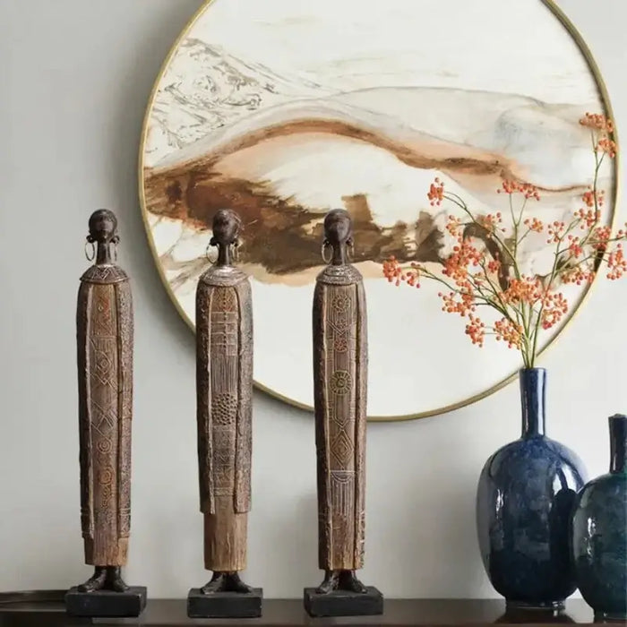 Elegant African style woman statues with intricate patterns, displayed in front of a decorative art piece and vase with branches.