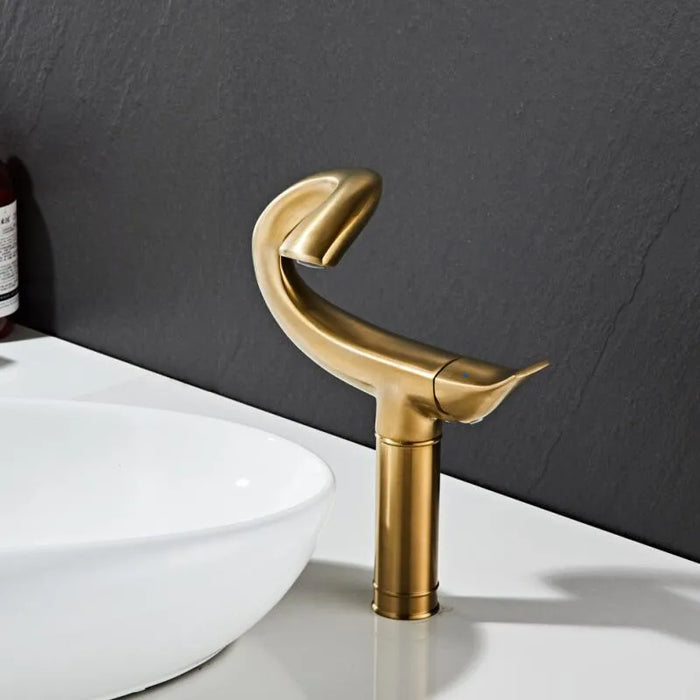 Elegant flamingo design deck-mount basin faucet in brushed gold finish next to a white ceramic sink in a modern bathroom setting.