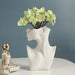 Modern Nordic ceramic vase with woman body model design, featuring green flowers, beside a small metallic sculpture.