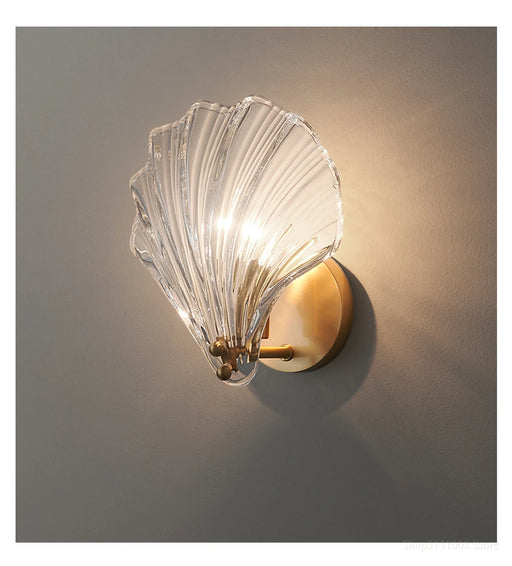 Modern luxury brass shell wall lamp with clear glass shade illuminating a wall, showcasing elegant design and craftsmanship.