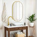 Oval wall-mounted mirror with gold aluminum frame above a wooden vanity in a modern bathroom setting.