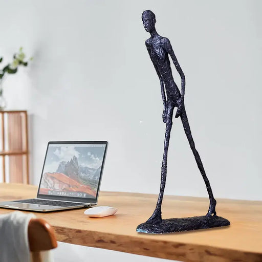 Bronze Giacometti-inspired Walking Man Sculpture on wooden desk near laptop and mouse, showcasing modern abstract art style.