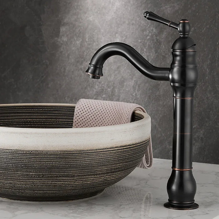 Antique brass long neck basin faucet with ceramic spool and single handle, paired with a textured stone bowl on a marble countertop.