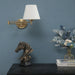 Gold folding swing arm wall lamp with white fabric shade above decorative table with horse sculpture and flowers.